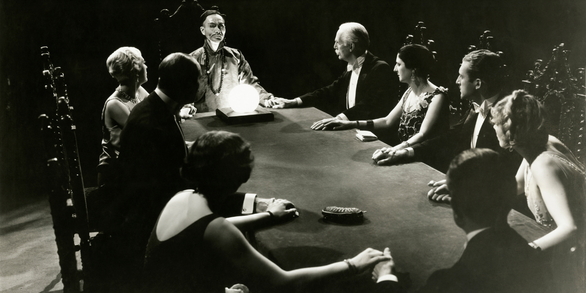 A group of people in 1920’s fashion gathers around a table to perform a seance.