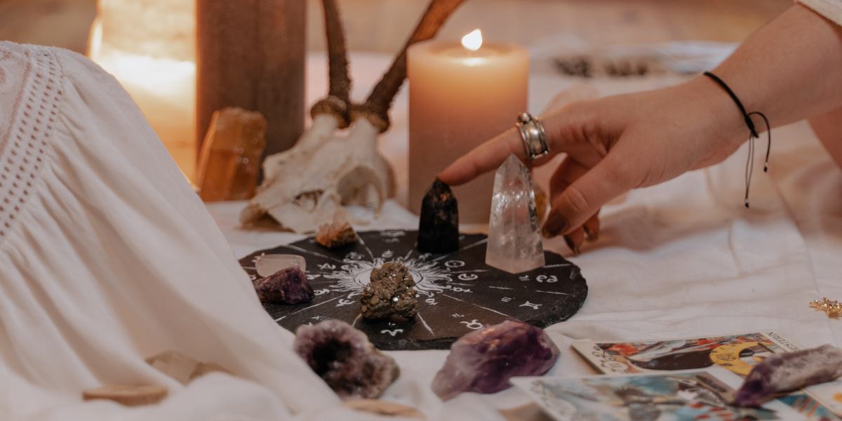 A hand touches crystals laid out on a board of zodiac symbols with tarot cards nearby.